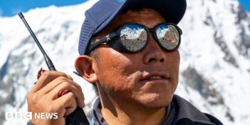 A Nepalese man wearing a cap and sunglasses, holding a walkie-talkie with snowy mountains behind him